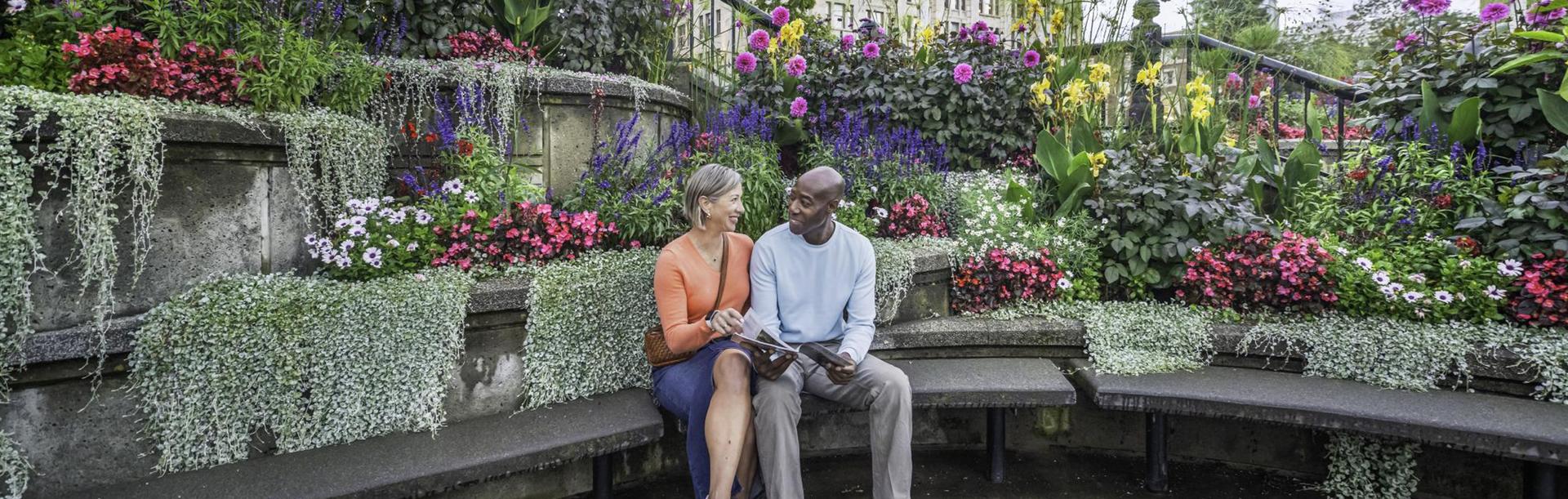 A couple reads the Victoria Vacation Guide while backdropped by flowers along the Upper Causeway of Victoria, BC's Inner Harbour