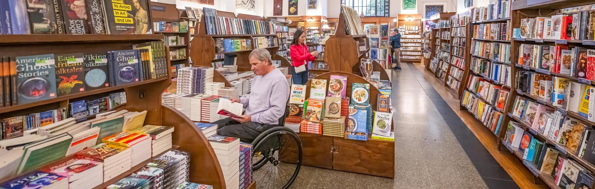 A man in a wheelchair and a woman in a pink coat shops for books in Munro's Books, a bookstore in Victoria, BC