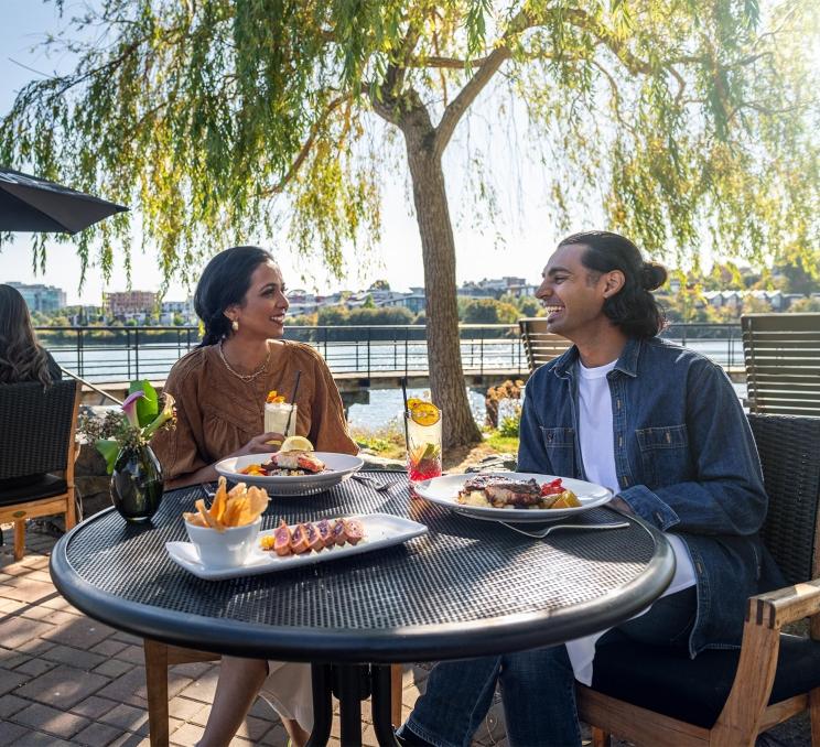 A young couple dines on fresh seafood on the patio at Glo Restaurant + Lounge, beneath a willow tree, along the Upper Harbour in Victoria, BC