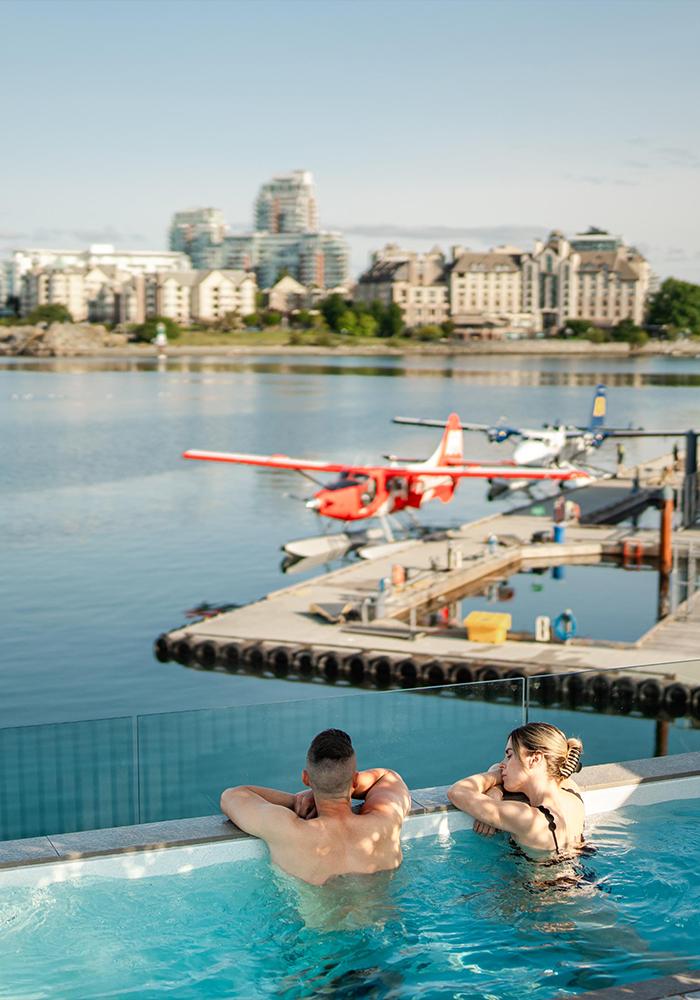 A couple soaks in a hot tub overlooking a red seaplane docked on Victoria, BC's Inner Harbour