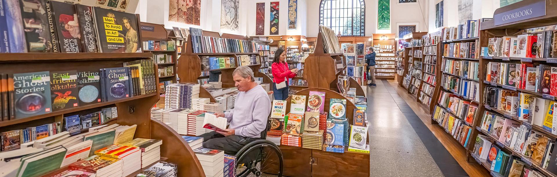 A man in a wheelchair and a woman in a red coat shop for books amongst the aisles at Munro's Books, a bookstore in a heritage buildings, in Victoria, BC