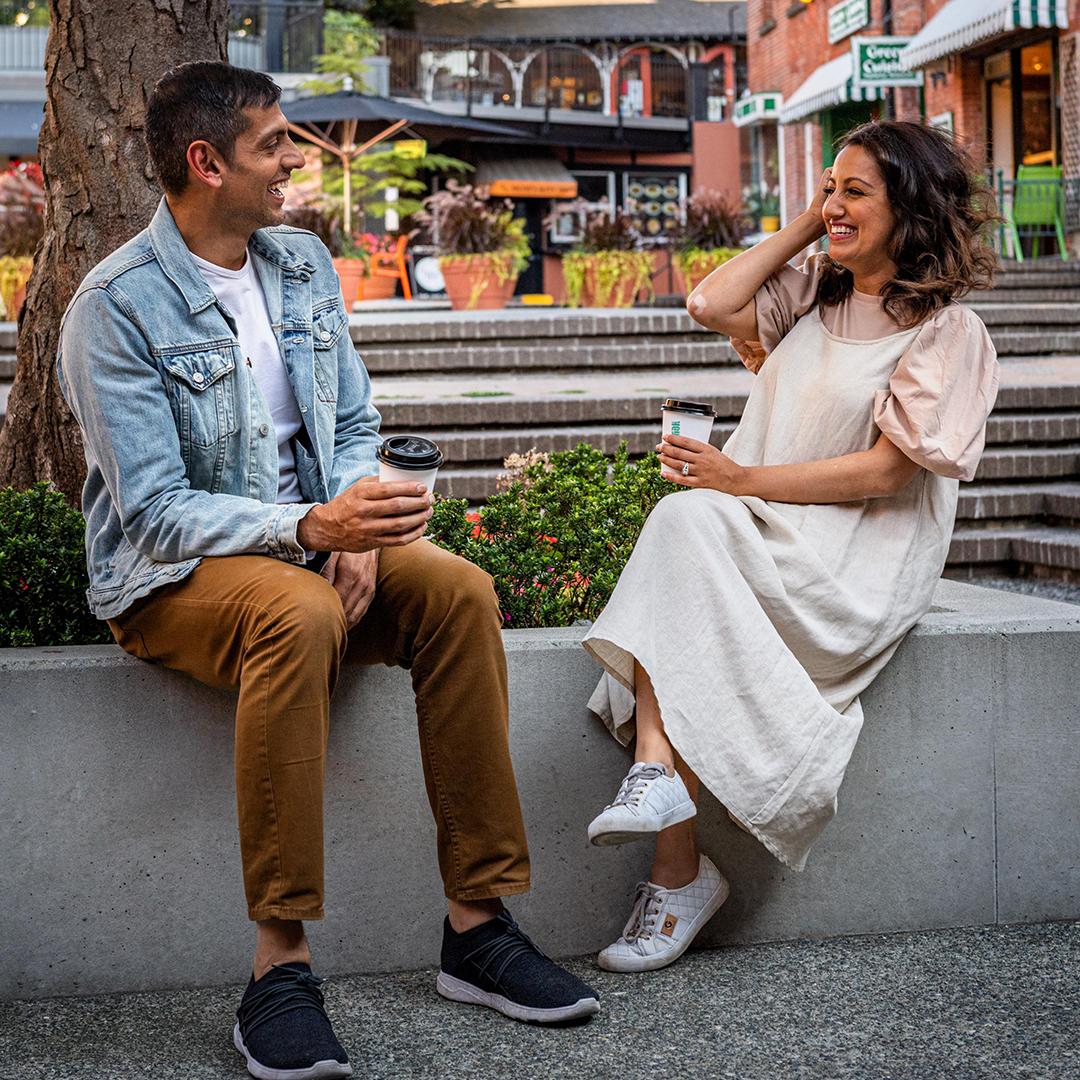 A couple, a man dressed in a denim jacket and woman in a cream dress, enjoy a coffee in the courtyard of Market Square in Victoria, BC