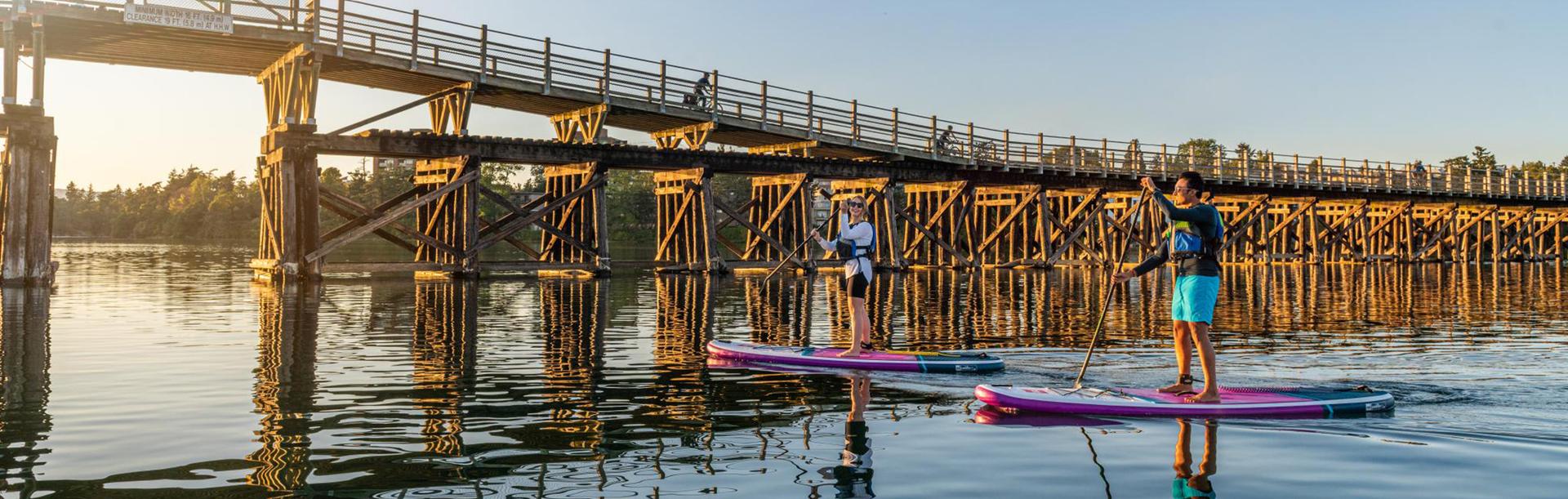 Two paddleboarders, a man in blue shorts, and a woman in a white shirt, paddle purple paddleboards across the Gorge Waterway at sunset, backdropped by the Selkirk Trestle, in Victoria, BC