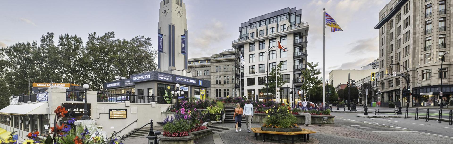 A woman in an orange sweater and a man in a blue sweater walk hand-in-hand along the Upper Causeway of the Inner Harbour in downtown Victoria, BC