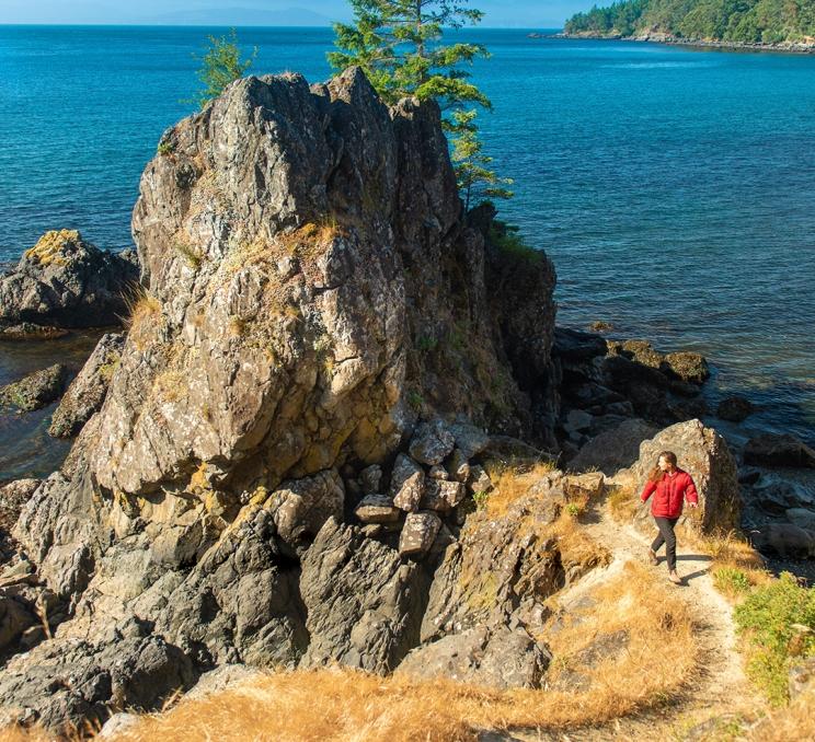A woman in a red jacket hikes along a seaside trail past a sea stack at Aylard Farm in East Sooke Regional Park in Greater Victoria, BC